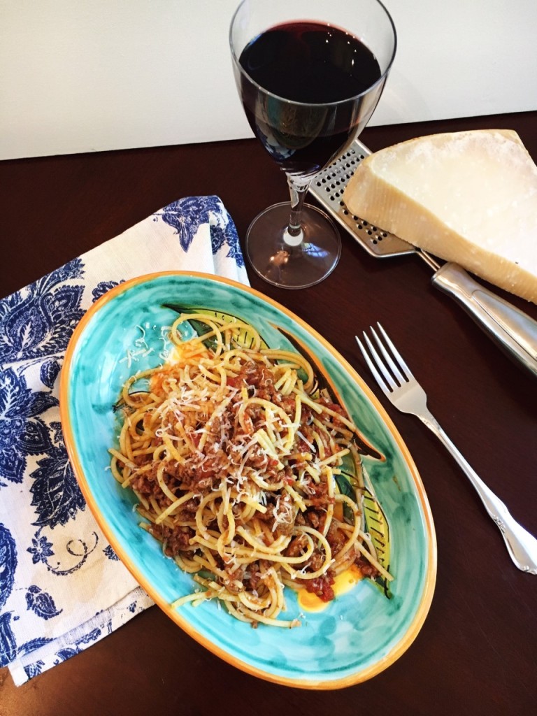 An oval plate of spaghetti with bolognese. A glass of wine and parmesan cheese at the side.